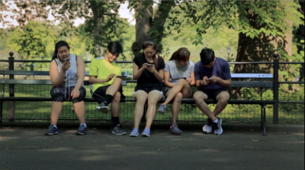 Film still: a park bench of people staring at their phones